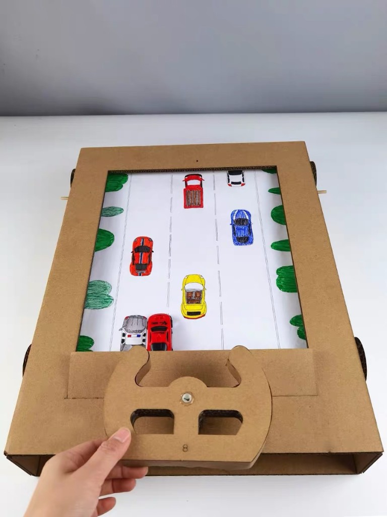 Handmade cardboard driving game with a paper road, toy cars, and a cardboard steering wheel