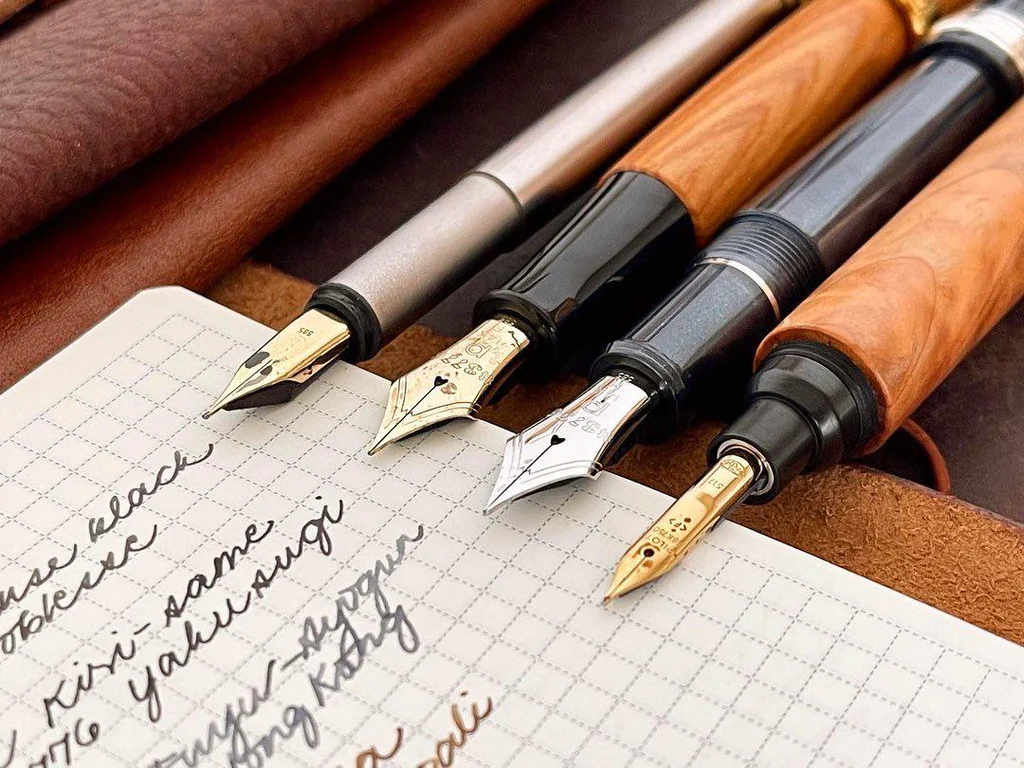 Four fountain pens on grid paper with cursive handwriting over a brown leather surface